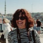 A joyful woman with curly hair and sunglasses, holding a camera outdoors on a sunny day.